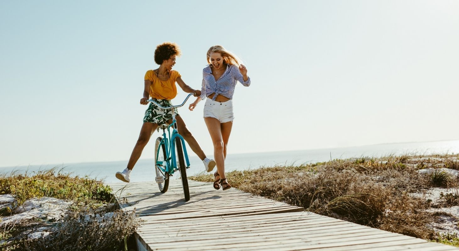 a woman riding a bike on a boardwalk by the beach while another woman is running alongside her