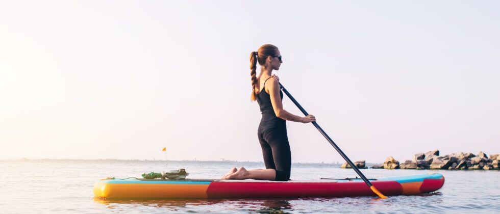 a woman on her knees on a paddle board close to shore on a cloudless day