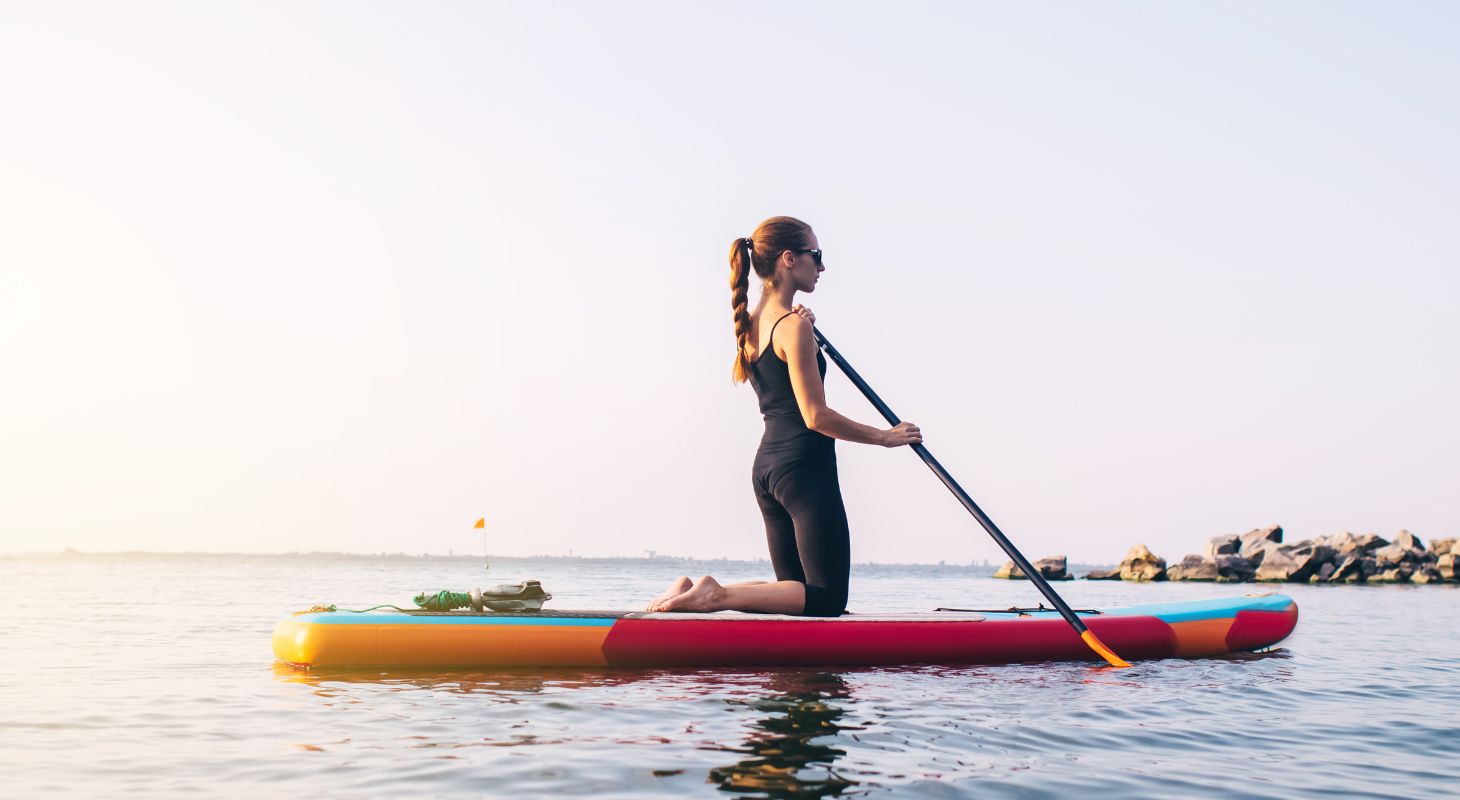 a woman on her knees on a paddle board close to shore on a cloudless day