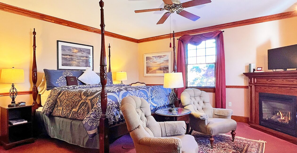A wooden 4-poster bed with cornflower blue and cream bedding with time period cream chairs and burgundy carpet.