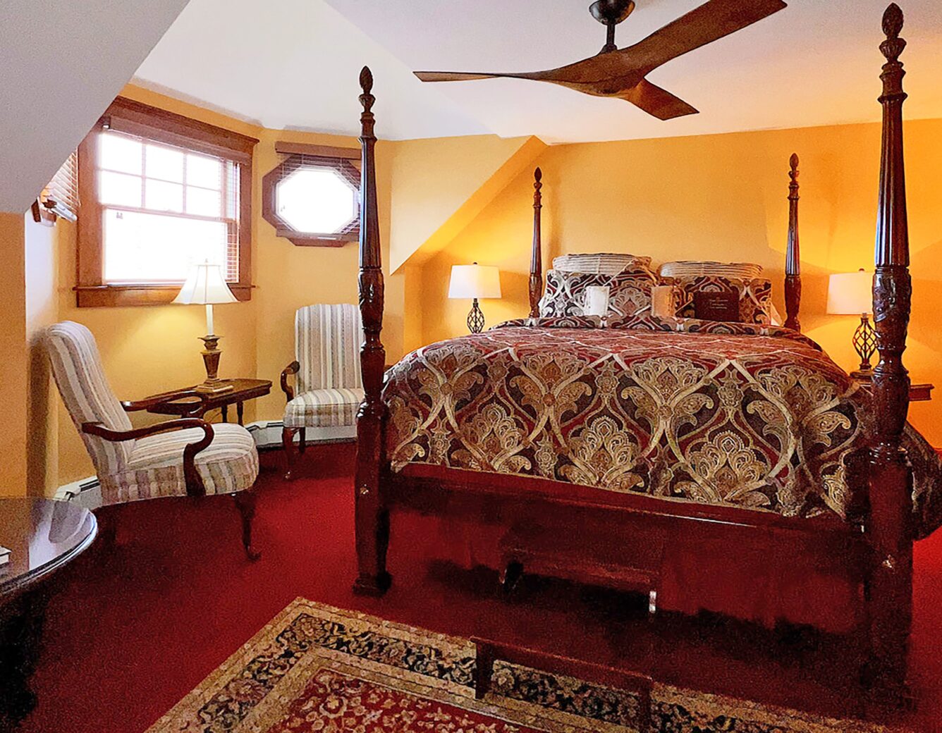 A beautiful wooden 4-poster bed with burgundy and gold bedding and time period chairs, lamps and rug