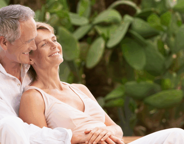 An older husband and wife with white hair recline and cuddle together with white clothing in front of greenery
