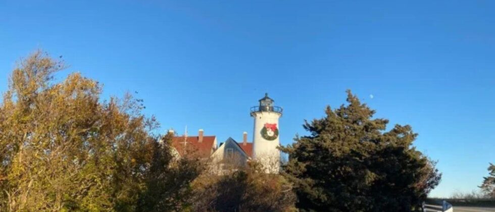 Lighthouse decorated with a holiday wreath surrounded by trees beneath a clear blue sky.