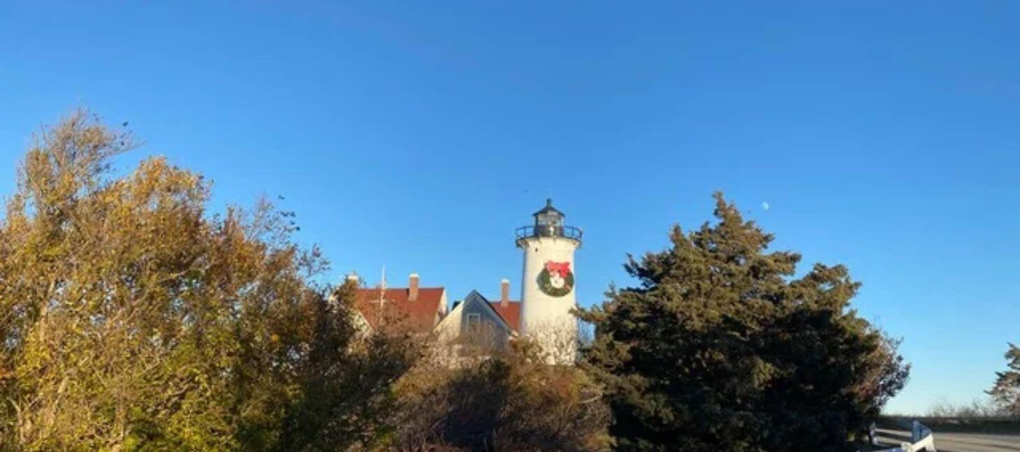 Lighthouse decorated with a holiday wreath surrounded by trees beneath a clear blue sky.